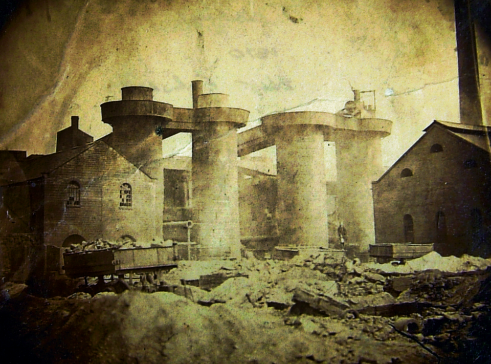 A sepia-toned photo shows a ruined industrial site with large cylindrical structures, debris on the ground, and damaged brick buildings, evoking a sense of historical destruction.